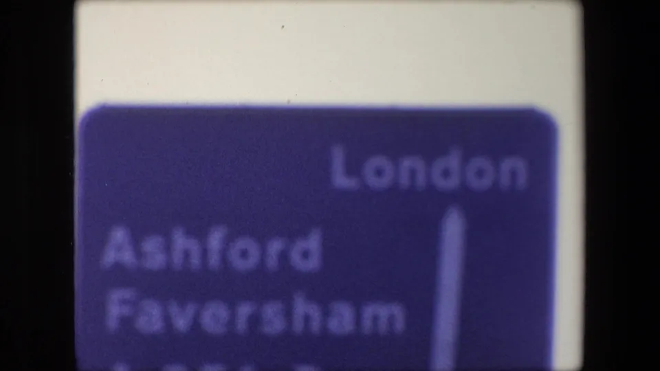Soft 8mm frame from a 1967 British travel clip showing a blue road sign with London, Ashford, and Faversham lettering partly visible.