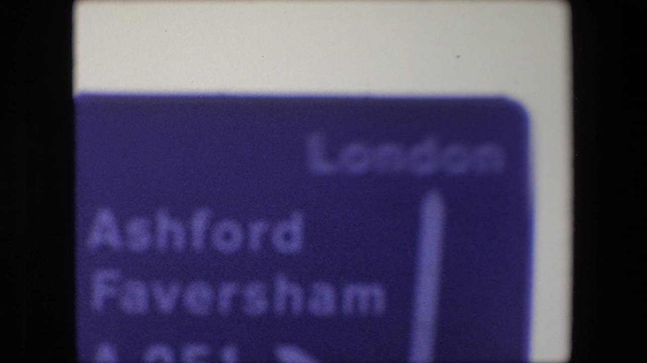 Blurred blue road sign with London and Ashford Faversham text beside a vertical arrow.