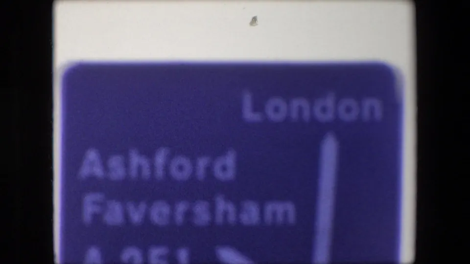 1967 8mm frame showing a blue British road sign with London, Ashford, and Faversham text, cropped by the film frame.