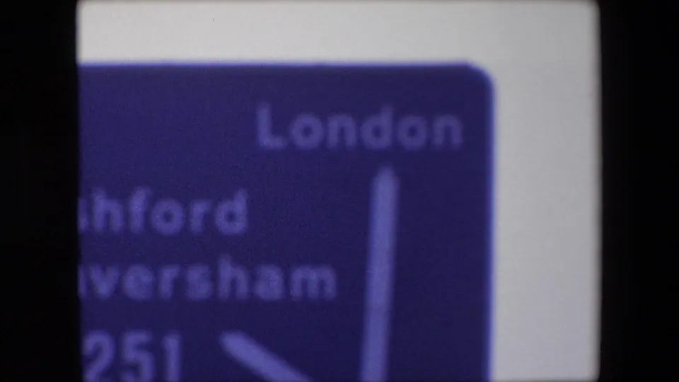 Cropped 8mm frame of a 1967 England directional sign with London, Ashford, Faversham, A251, and arrow markings visible in soft focus.