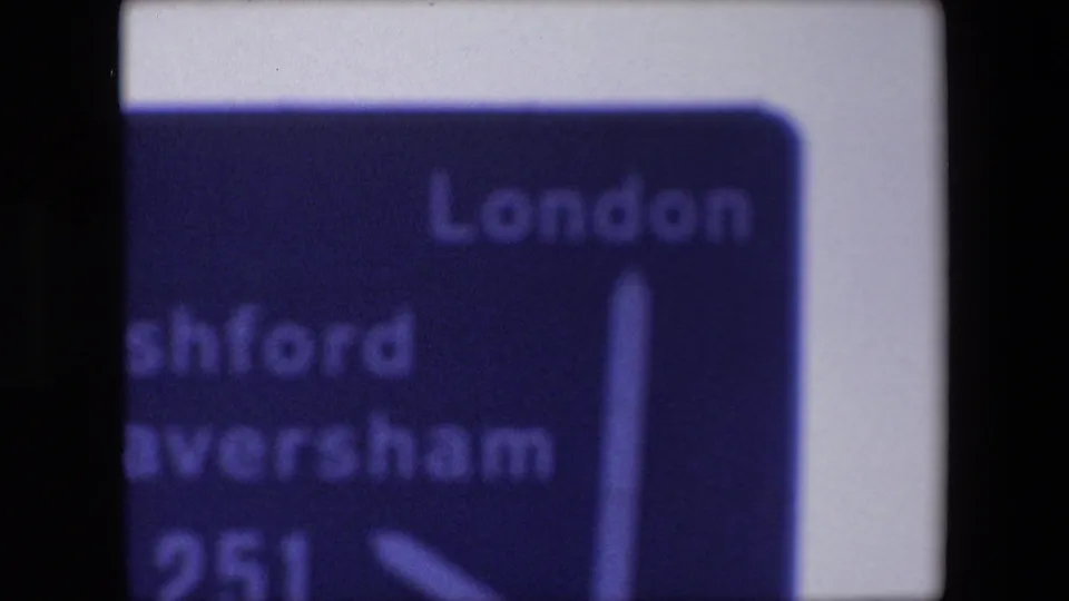 Late frame from the 8mm clip showing the blue 1967 British road sign cropped around London, Ashford, Faversham, A251, and white direction arrows.