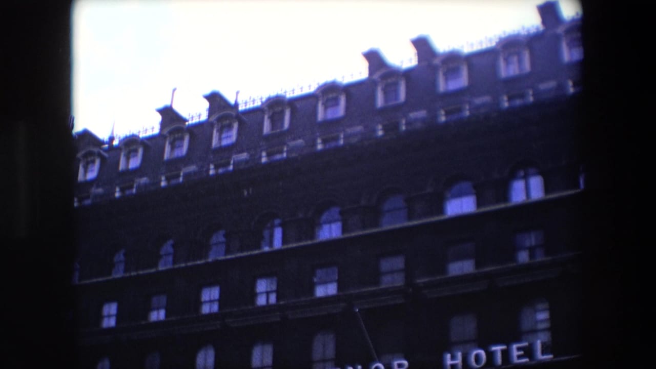1967 London home-movie frame showing the upper stories of a dark brick building with arched windows, roof dormers, and partial hotel lettering at the bottom edge.