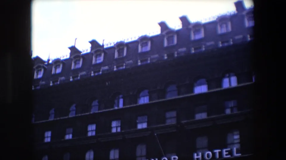1967 London home-movie frame showing the upper stories of a dark brick building with arched windows, roof dormers, and partial hotel lettering at the bottom edge.