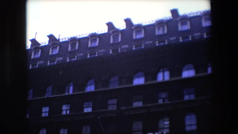 1967 London exterior frame with a low-angle view of a dark brick multi-story facade, repeated windows, dormers, and heavy film vignetting at the edges.