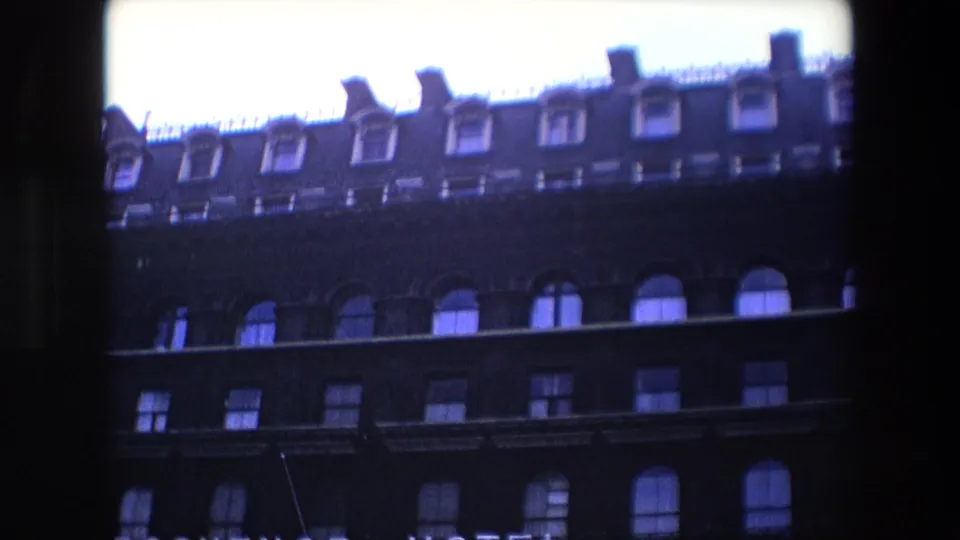 1967 London 8mm frame showing a historic-looking building facade with rows of arched windows, roof dormers, overexposed sky, and a partial hotel sign.
