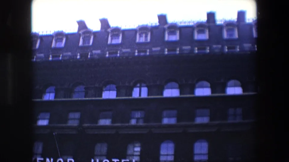 1967 London home-movie frame capturing the building's upper stories, repeated arched windows, dormers, a bright sky, and cropped hotel lettering at the lower edge.