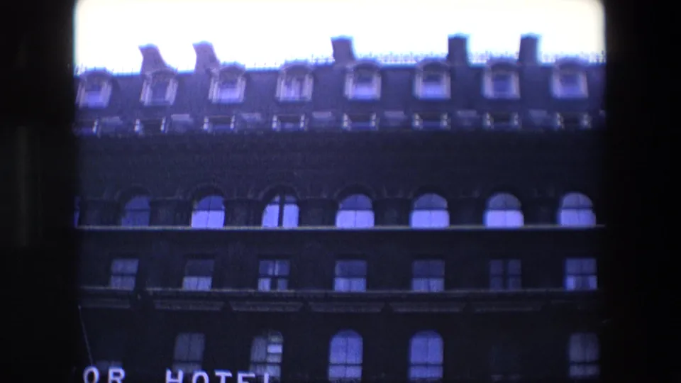 1967 London 8mm frame showing a dark multi-story hotel facade from below, with arched upper windows, rectangular lower windows, roof dormers, and part of an illuminated HOTEL sign.
