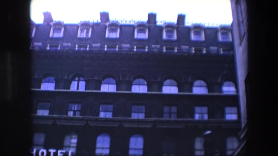 1967 London 8mm frame of the hotel exterior, showing the upper roofline, dormer windows, arched window row, and partial illuminated HOTEL lettering near the lower edge.