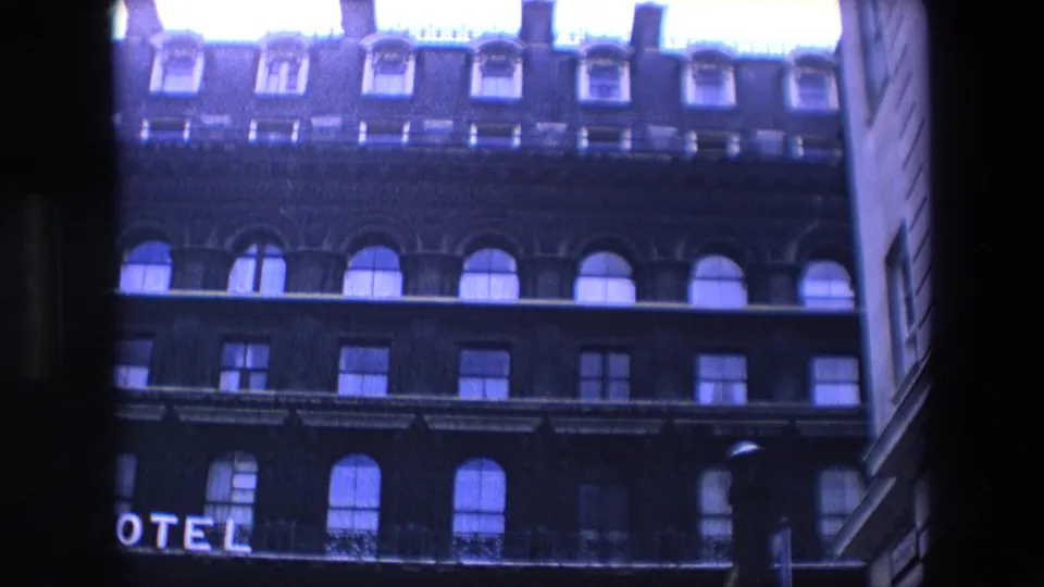 1967 London 8mm frame showing the hotel facade cropped lower in the view, with roof dormers, arched windows, rectangular windows, partial HOTEL letters, and a streetlamp at the right edge.