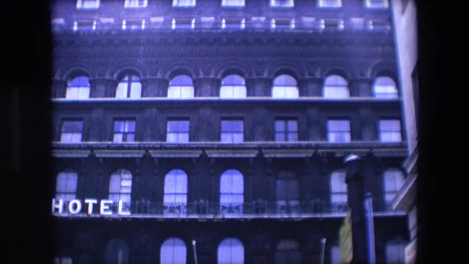 1967 London 8mm frame of the dark hotel exterior with a clearer partial HOTEL sign, balcony railing, arched windows, rectangular windows, and a streetlamp along the right side.