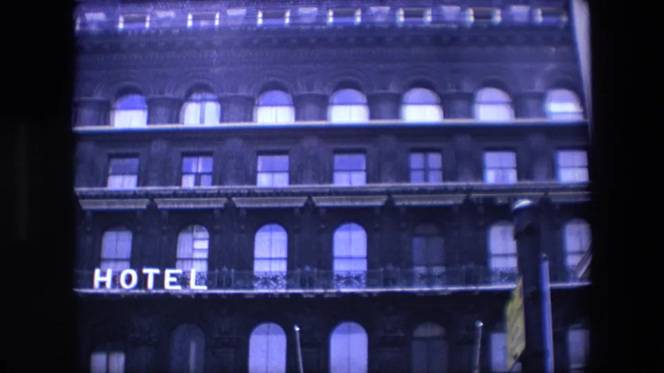 1967 London home-movie frame showing part of the Grosvenor Hotel facade with arched windows, balcony railings, and partial illuminated hotel lettering.