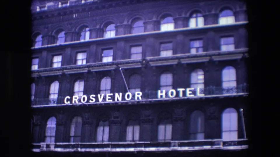 1967 London home-movie frame showing the Grosvenor Hotel name clearly lit across the brick facade, with arched windows and balcony railings visible above and below.
