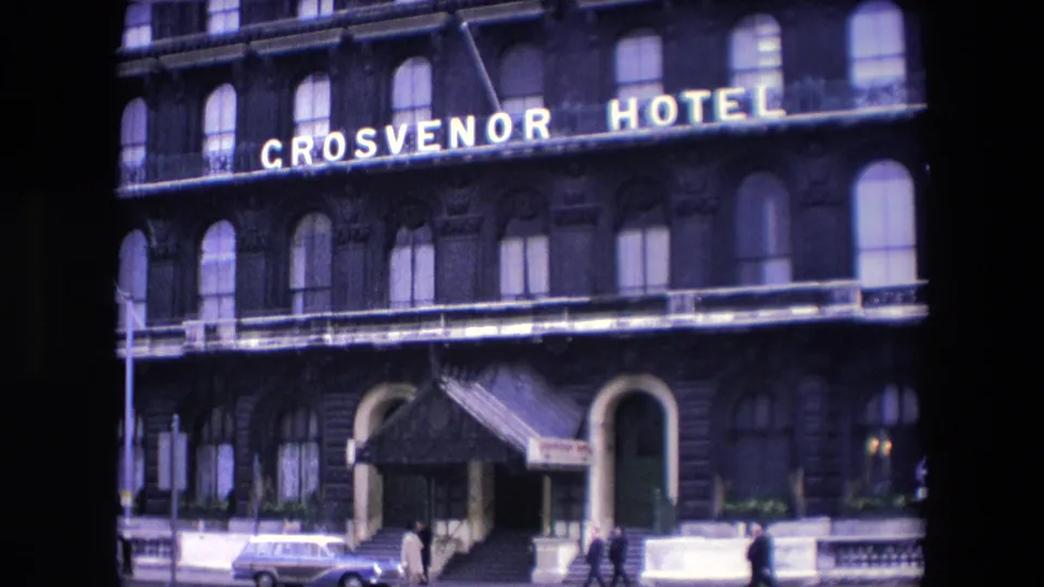 1967 London 8mm frame showing the Grosvenor Hotel entrance with steps and canopy, pedestrians on the pavement, a street lamp, and a parked estate car.