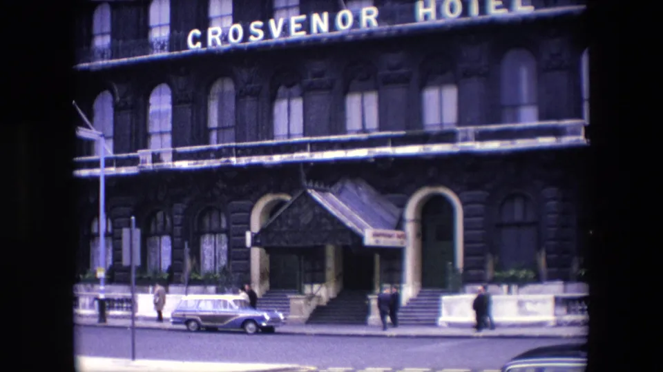 Wider 1967 London frame of the Grosvenor Hotel exterior, showing the entrance canopy, arched doorway, pedestrians, a parked estate car, and a vehicle partly entering the foreground.