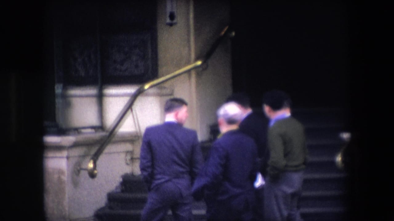 In 1967 London home-movie footage, several adult men in suits are seen from behind as they climb stone steps beside brass handrails toward a dark entrance.