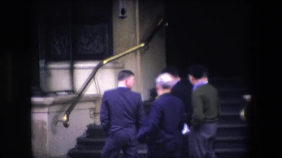 In 1967 London home-movie footage, several adult men in suits are seen from behind as they climb stone steps beside brass handrails toward a dark entrance.
