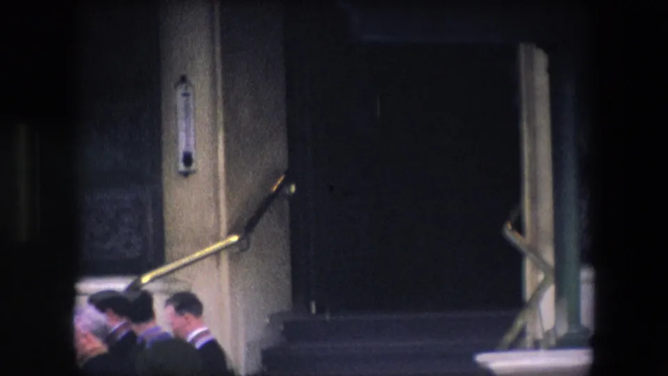 A small group of adult men in suits stands or moves near the base of stone steps at a formal London building entrance, with brass railings and heavy masonry visible.