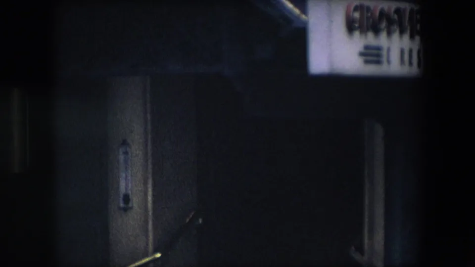 A dim, close view of the building entrance shows stonework, a brass rail, and partial signage near the top edge, with the handheld 8mm framing obscuring details.