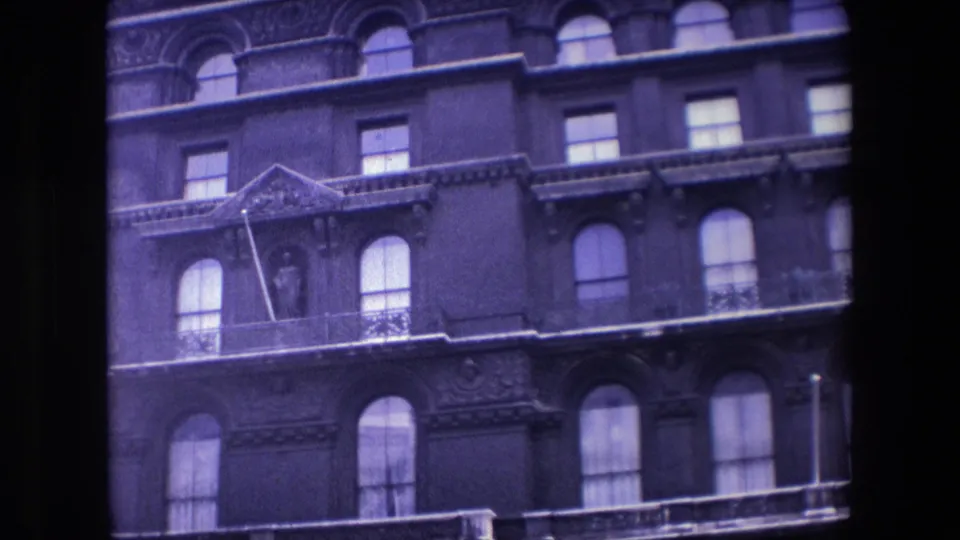1967 London home-movie frame with a closer handheld view of the ornate building facade, including arched windows, a balcony edge, and a statue set into the wall.