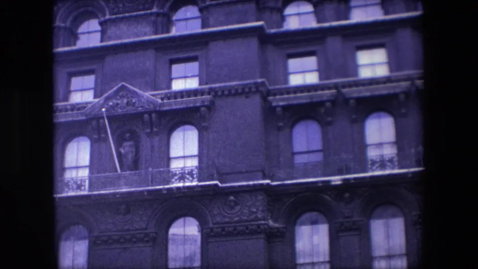 1967 London 8mm frame showing the camera moving across an ornate facade with a triangular pediment, balcony railing, arched windows, and soft purple-blue color shift.