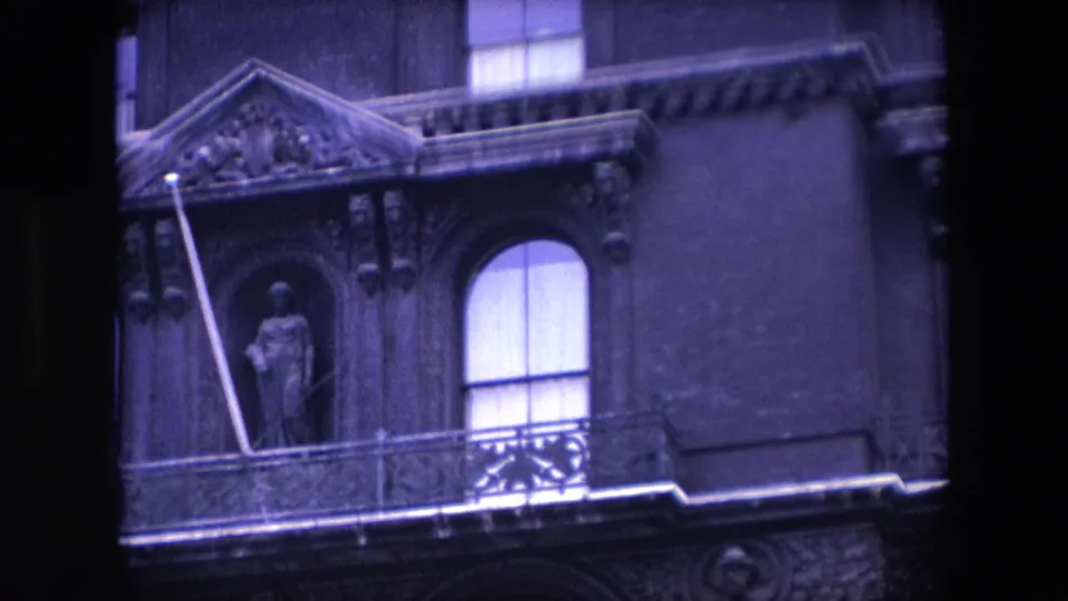 1967 London home-movie frame showing an ornate church facade with a curtained arched window, balcony rail, carved stone decoration, and statue in a niche.