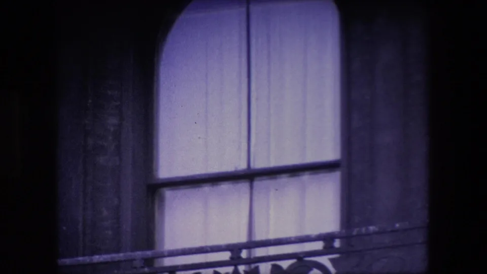 Close 8mm view of an arched curtained window and balcony railing on a dark stone London building, supported by the clip metadata.