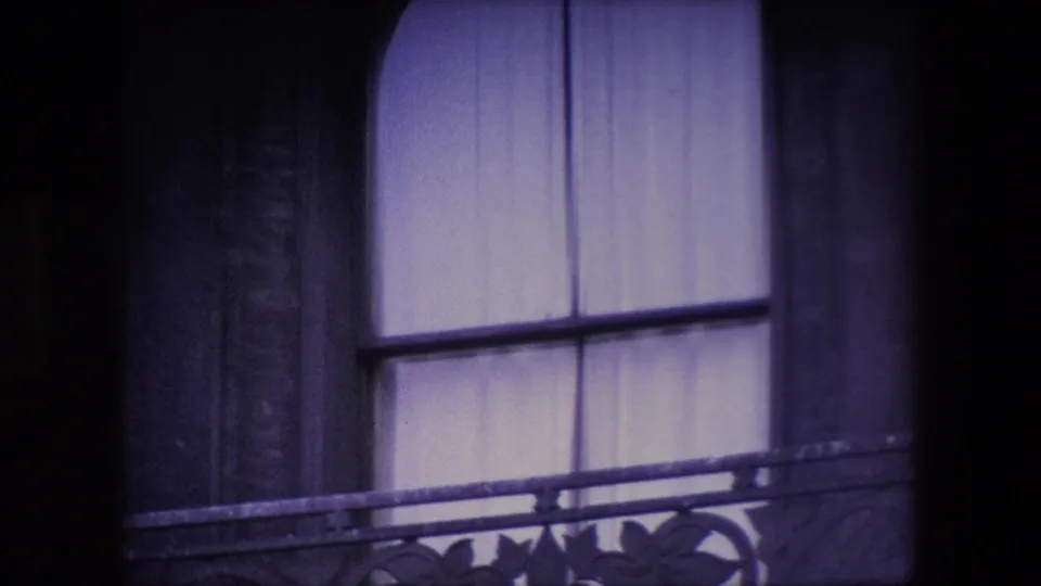 Shadowed close view of an arched window with pale curtains and decorative railing, matching the 1967 London home-movie setting supplied in metadata.