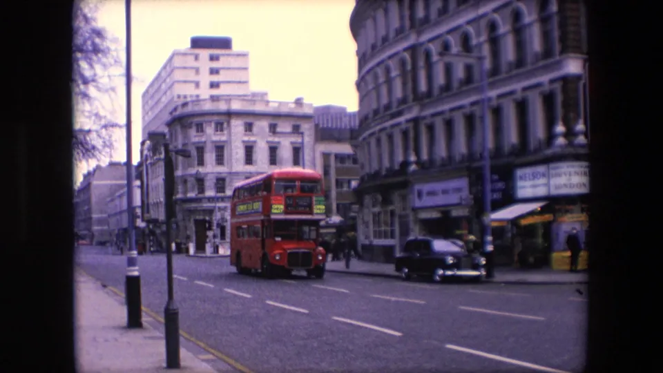 1967 London street view showing a red double-decker bus passing historic buildings, a black taxi, pedestrians, and visible shopfront signage.