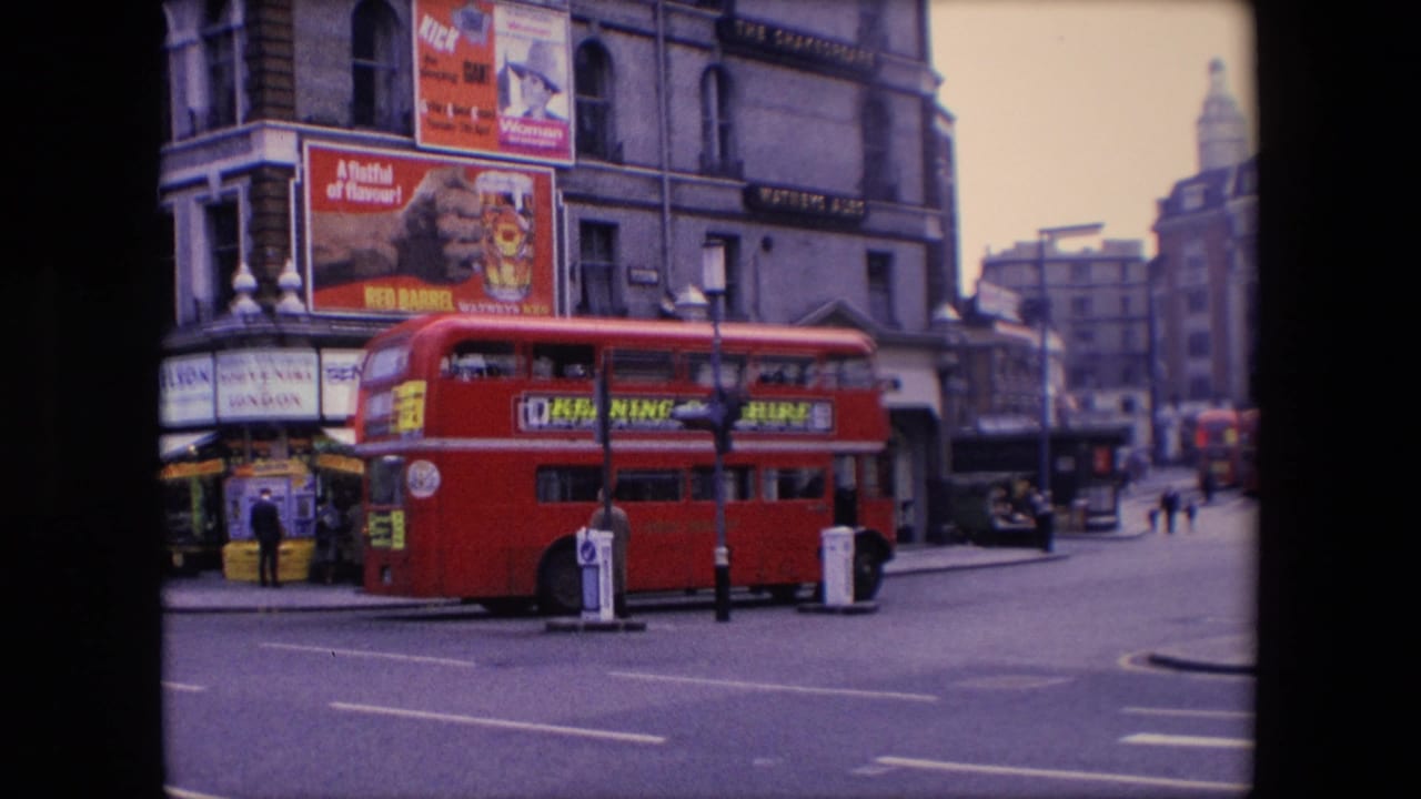 Red double-decker bus turning at a London street corner with shopfronts and poster advertisements behind it.