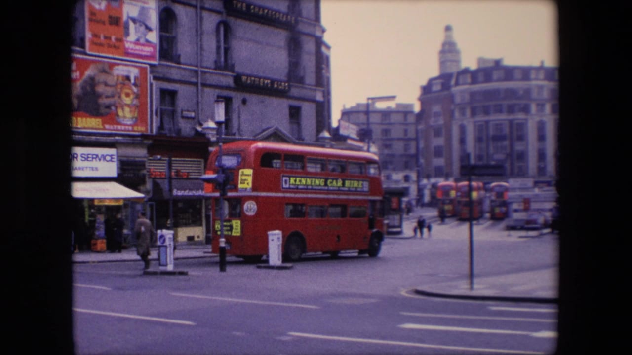 Red double-decker bus beside shopfronts at a broad intersection with pedestrians and another bus in the distance.