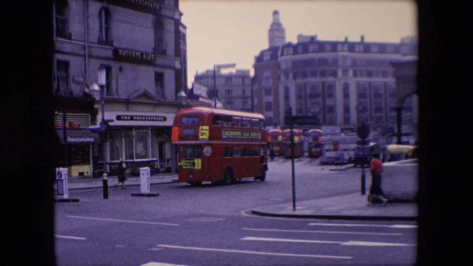 1967 London street view showing the rear of a red double-decker bus moving away, pedestrians near the corner, shopfronts, roadway markings, and city buildings.