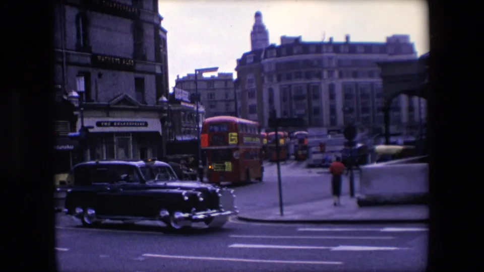 1967 London intersection with a black taxi crossing the foreground, a red double-decker bus beyond it, pedestrians, shopfront signage, and multi-storey buildings.