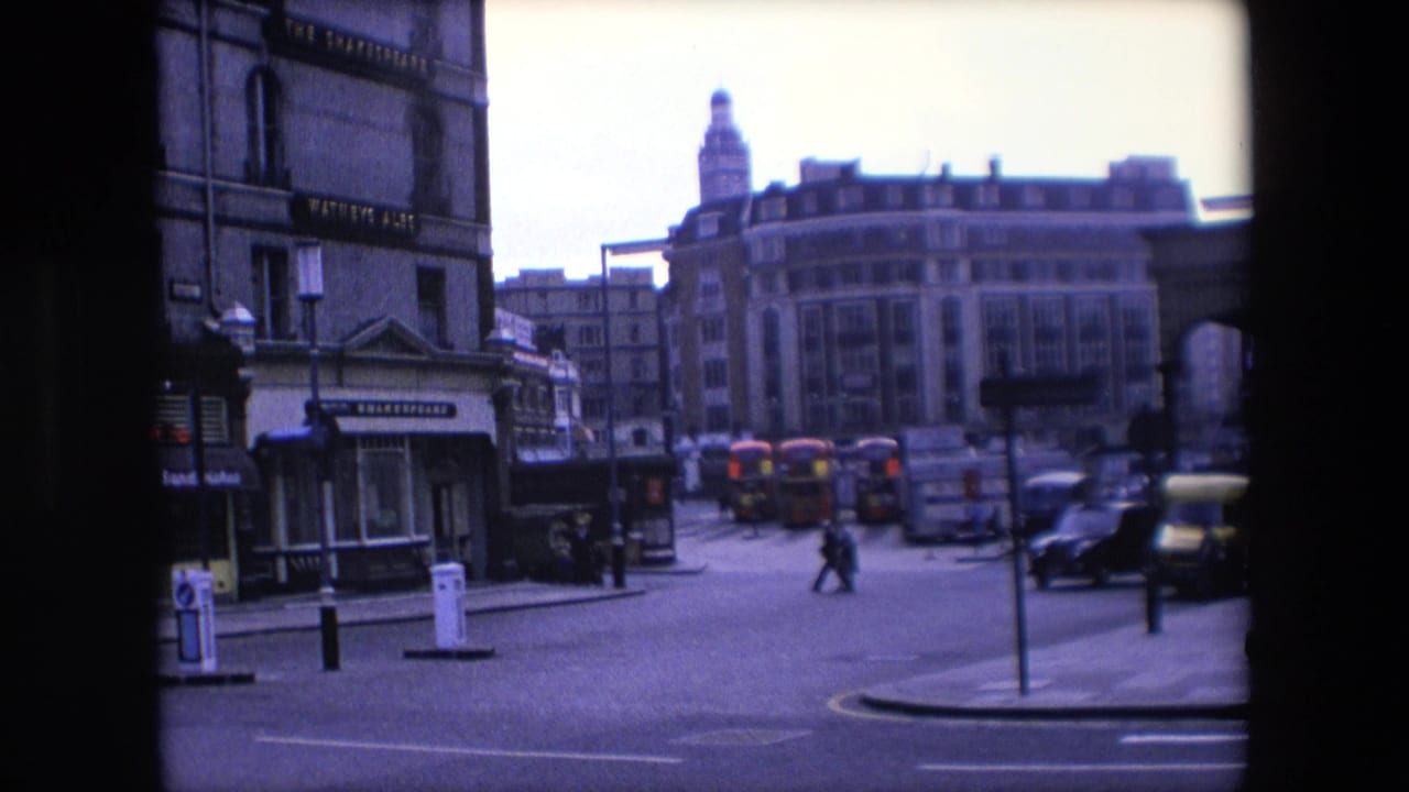 London street with pedestrians crossing, buses in the distance, and a dark car at right.