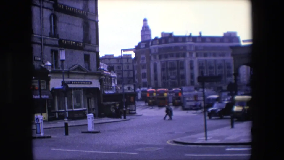 A 1967 London street view shows two pedestrians crossing an open roadway, red buses in the distance, building facades, bollards, and a dark vehicle near the curb.
