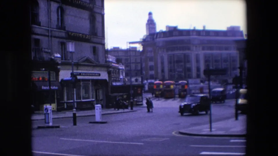 A black London taxi passes through the foreground while a pedestrian crosses the street, with red buses, bollards, shopfront signage, and multi-story buildings behind.