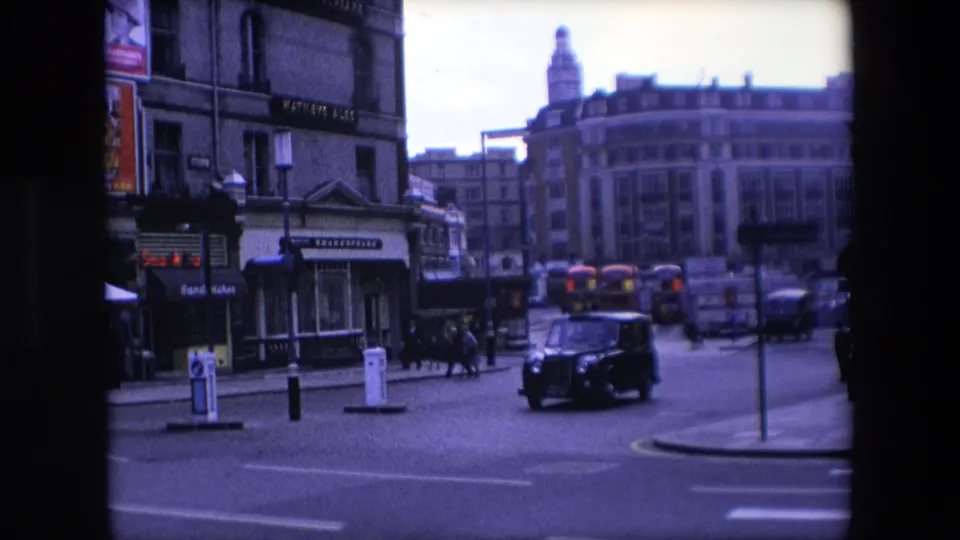 The later street frame shows a black taxi moving across the roadway, several pedestrians near the building fronts, distant red buses, street signage, and a Shakespeare sign on the left-side facade.