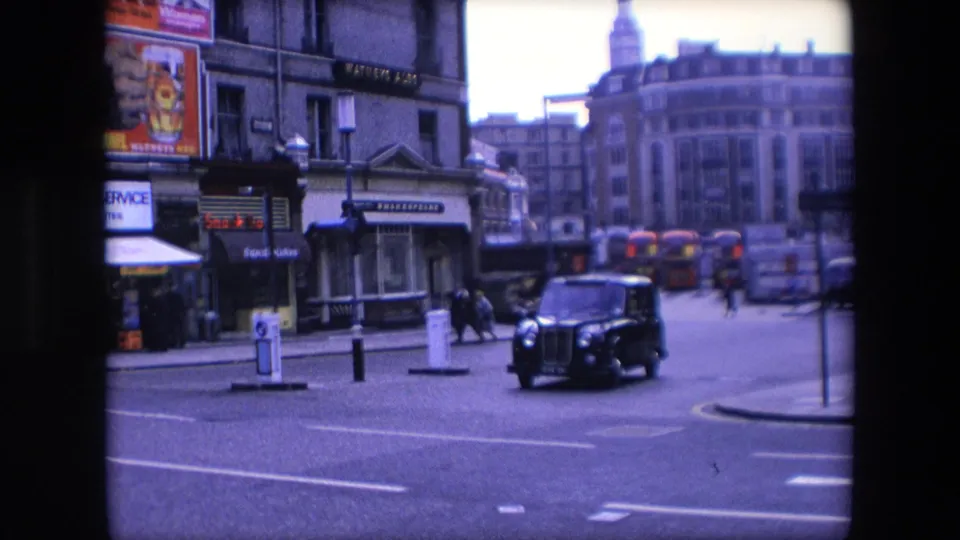 A black London taxi moves through a city street in 1967 London, with red double-decker buses, pedestrians, shop signs, and multi-storey buildings behind it.
