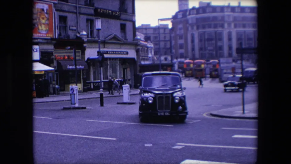The black London taxi turns at normal speed on a 1967 city street, with distant red buses, traffic signals, pedestrians, and commercial signs visible around the junction.