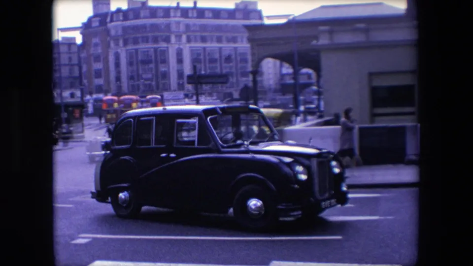 A side view shows a black London taxi passing close to the camera in 1967, with buses, pedestrians, an arched building detail, and city street markings behind it.