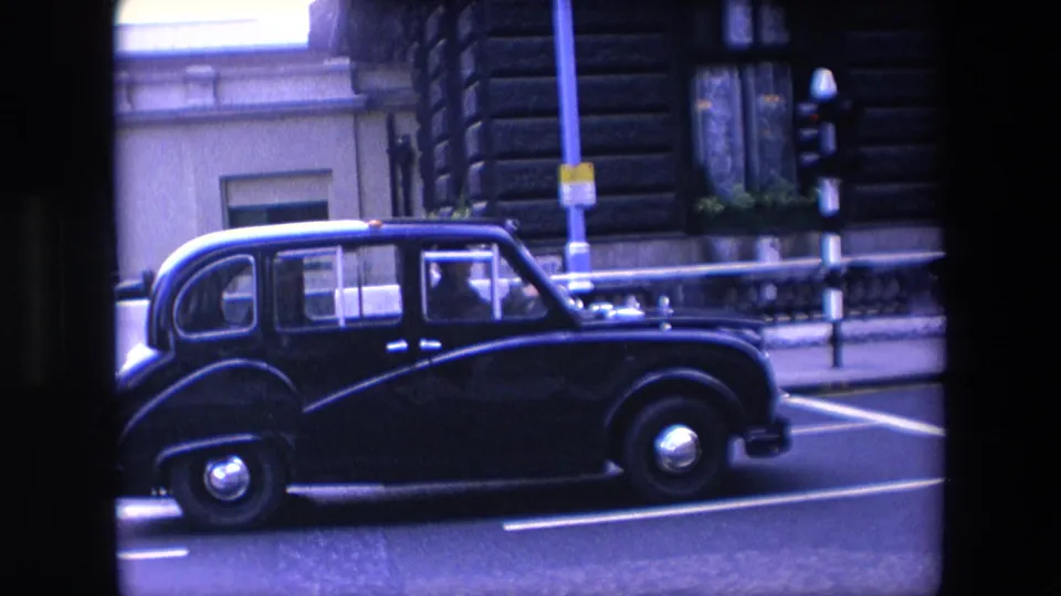 The taxi continues past the camera in profile on a London street, with a traffic light, lamp post, stone building facade, roadway lines, and pavement railings nearby.
