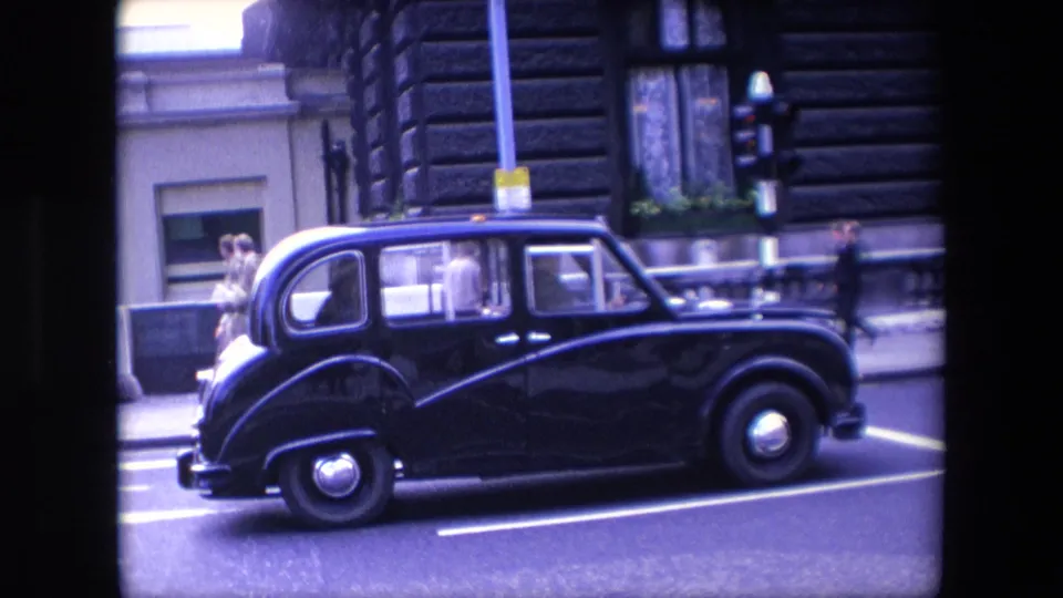 A classic black taxi waits at a traffic light on a London street in 1967, with pedestrians, stone building frontage, and painted road markings visible through the 8mm frame border.