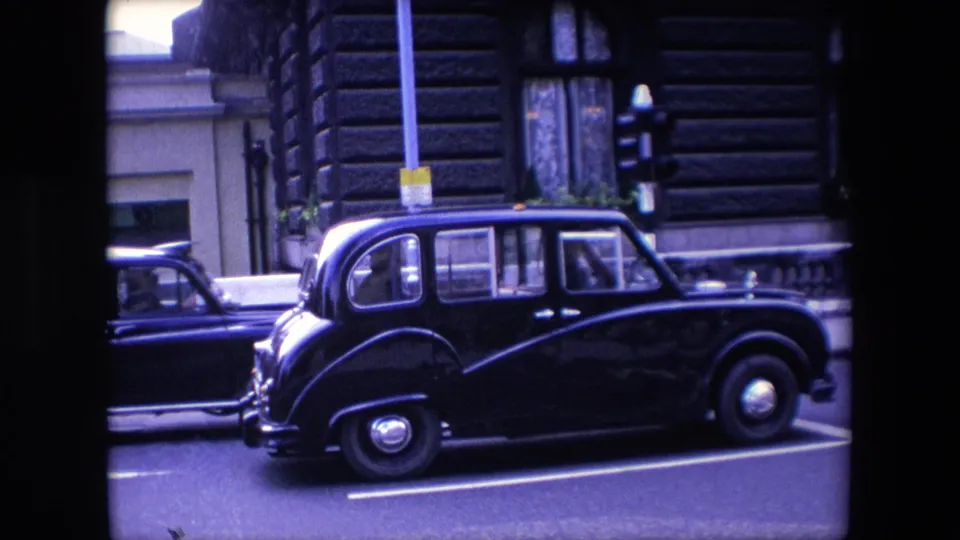 A black London taxi is seen in profile near a traffic signal in 1967, with another dark car partly visible behind it and building windows in the background.
