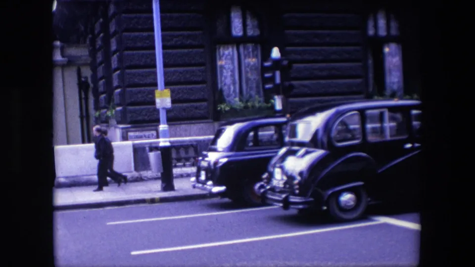 Two dark rounded vehicles, including a taxi-like car, move through a London street scene in 1967 as a pedestrian walks past barriers and a building facade.
