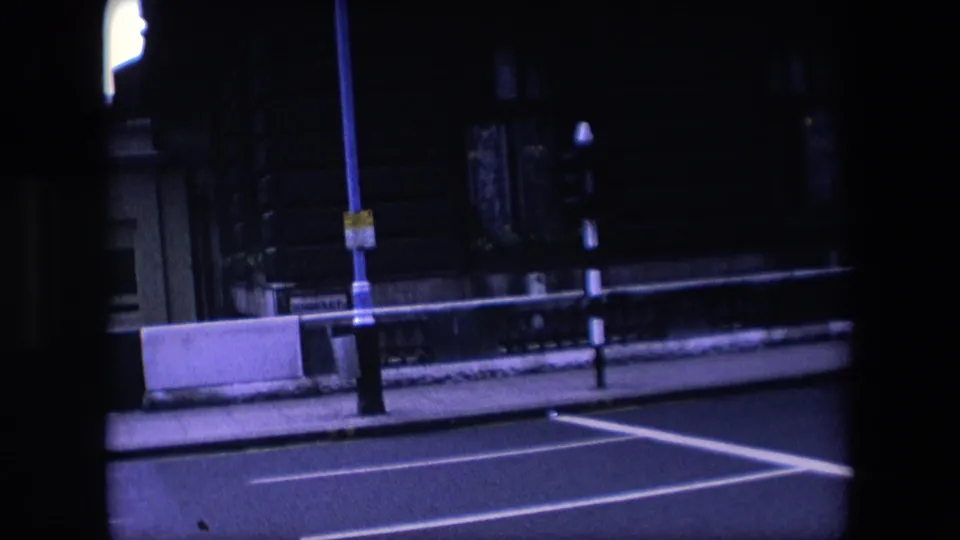 The street clears near the end of the 1967 London home-movie shot, leaving the traffic light, road markings, pavement barriers, and dark building frontage in view.