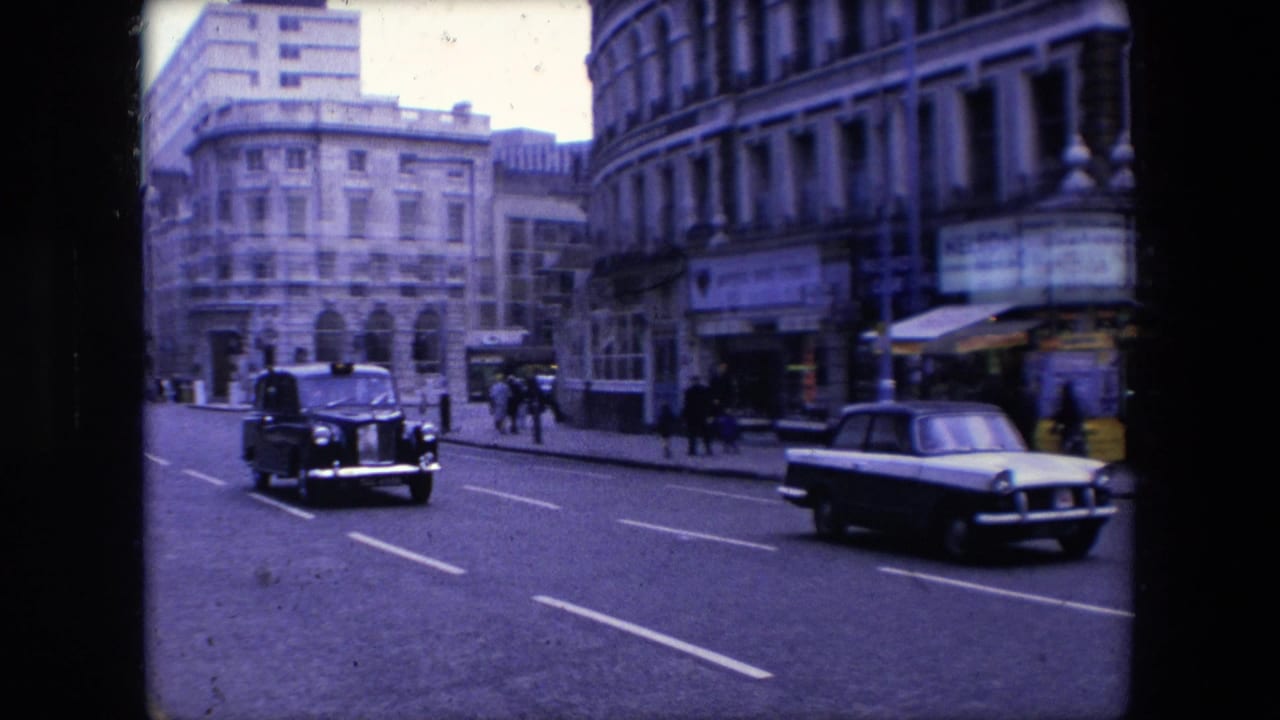 Black taxi and light-colored car driving along a city street beside stone buildings and pedestrians.