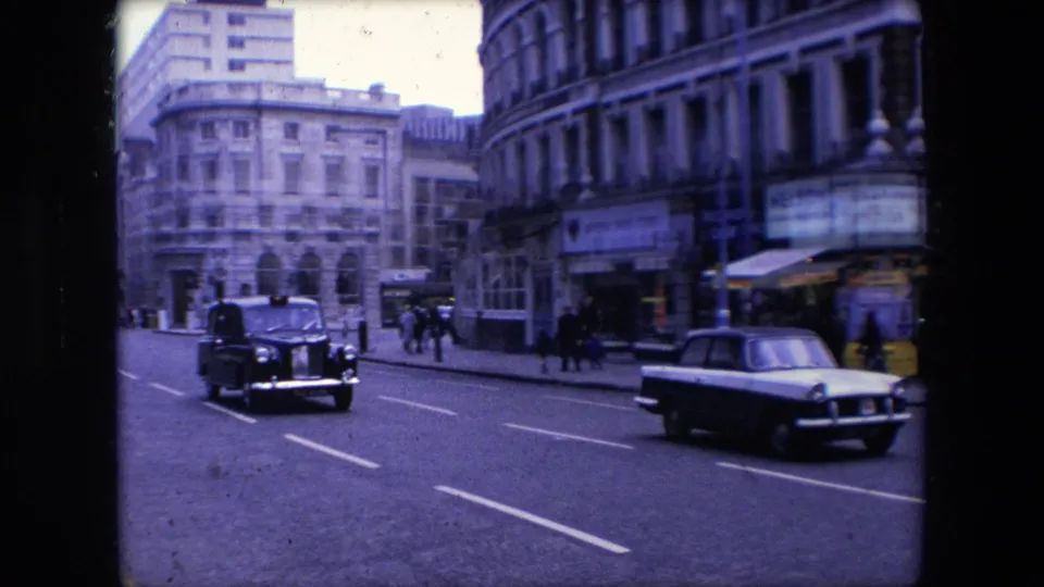 1967 London street view with a black taxi, a light-colored sedan, pedestrians on the pavement, and multi-story stone commercial buildings in the background.