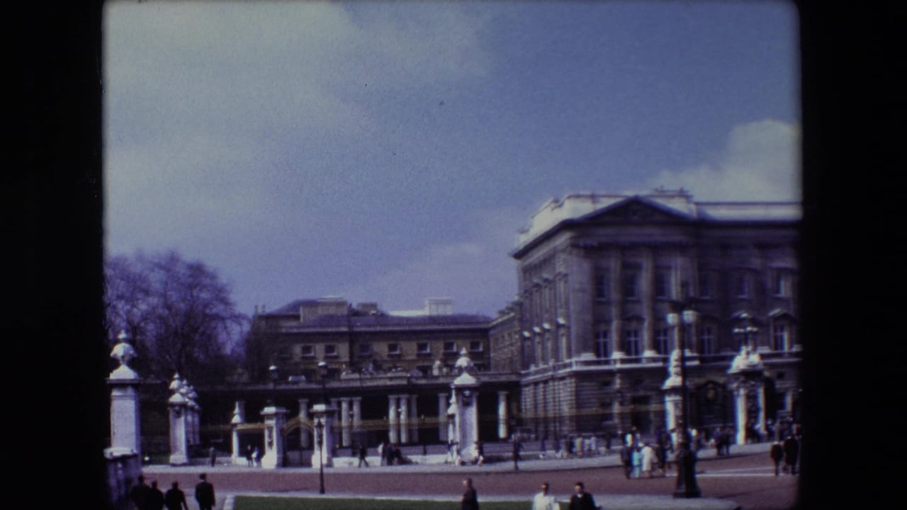 1967 London home-movie frame showing Buckingham Palace under a blue sky, with pedestrians near the palace gates and a broad forecourt in front.