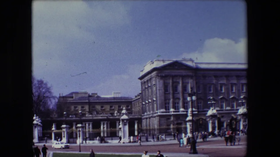 1967 London frame of Buckingham Palace with tourists and locals visible near the gate area and a light-colored car passing across the palace forecourt.