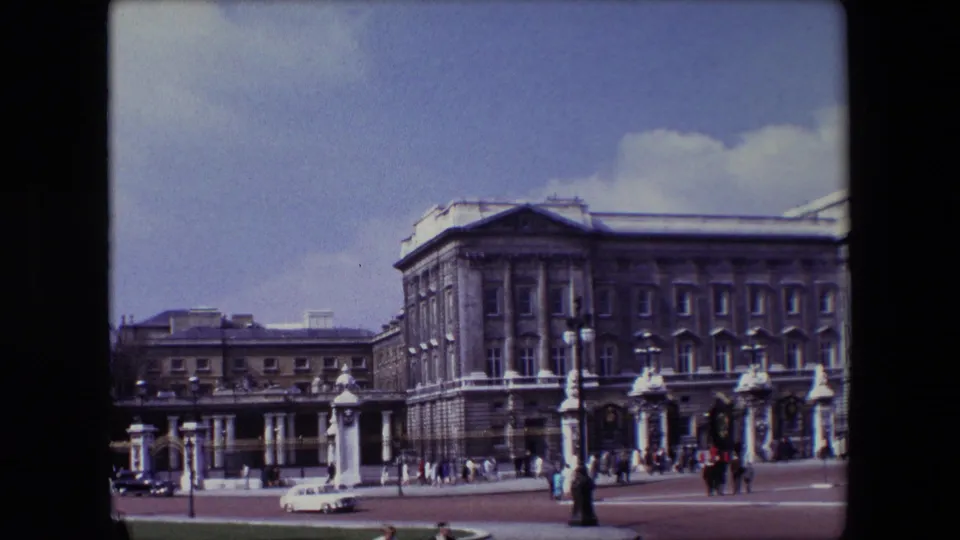 1967 London home-movie frame with Buckingham Palace filling the right side, pedestrians crossing the forecourt, and a pale car moving along the street.