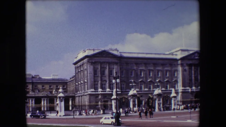 1967 London frame showing Buckingham Palace and its gate area with pedestrians crossing the forecourt as a white car and darker car pass through the scene.